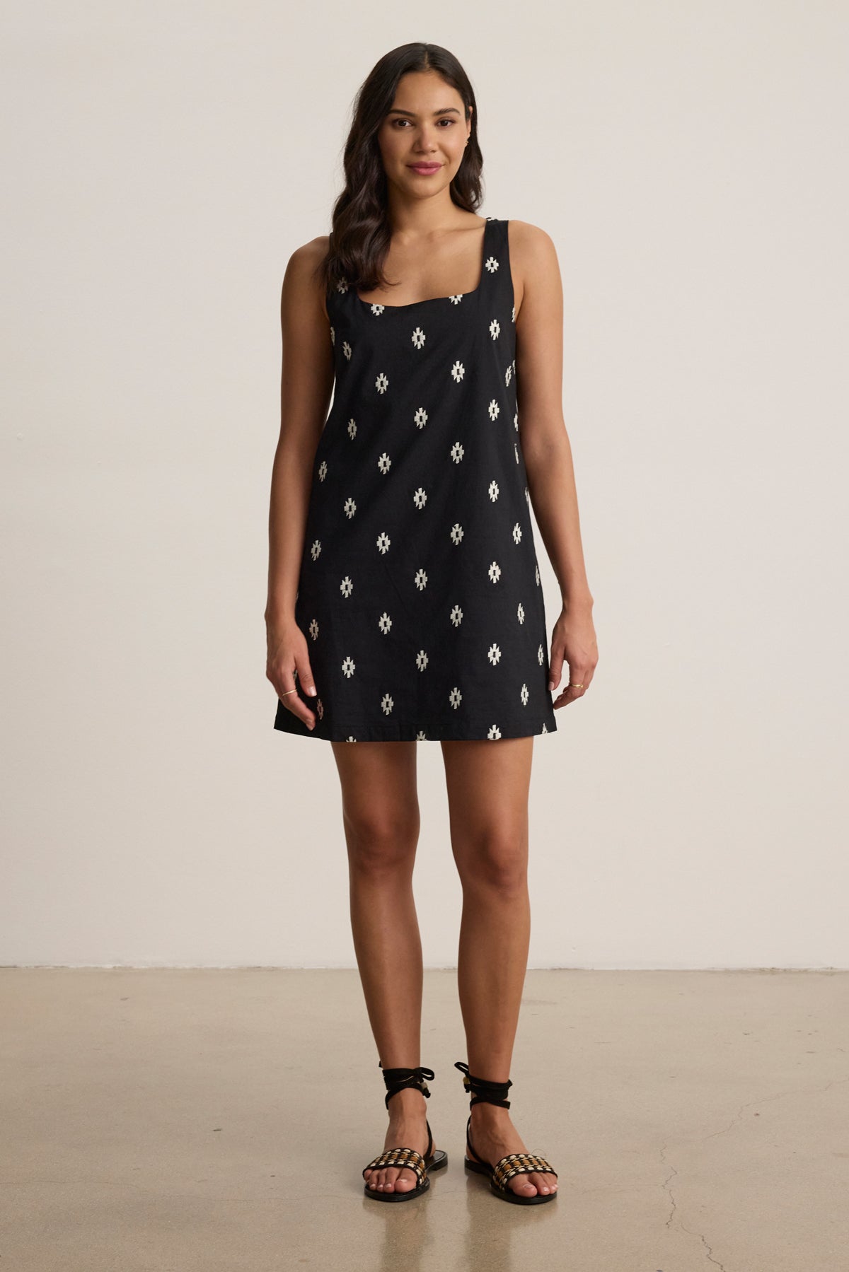 A woman stands indoors facing forward against a plain background, wearing the ELARA EMBROIDERED COTTON VOILE TANK MINI DRESS by Velvet by Graham & Spencer and strappy black sandals.