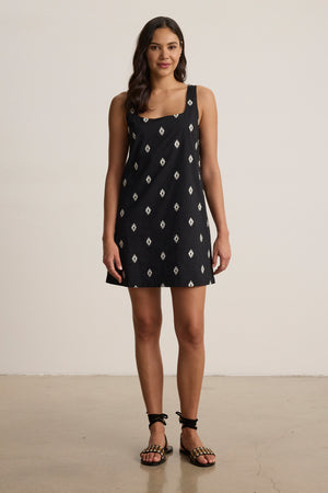 A woman stands indoors facing forward against a plain background, wearing the ELARA EMBROIDERED COTTON VOILE TANK MINI DRESS by Velvet by Graham & Spencer and strappy black sandals.