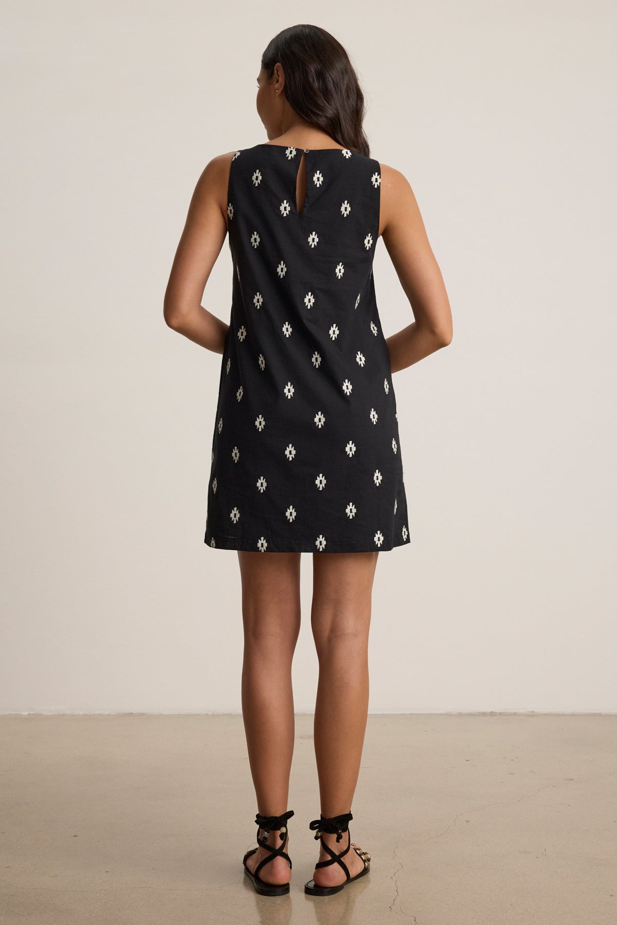 A woman stands indoors facing away, wearing the ELARA EMBROIDERED COTTON VOILE TANK MINI DRESS by Velvet by Graham & Spencer—a sleeveless black dress with a white geometric pattern—and black sandals on a plain floor.