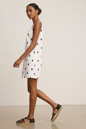 A woman wears the ELARA EMBROIDERED COTTON VOILE TANK MINI DRESS by Velvet by Graham & Spencer— a sleeveless white dress with black geometric patterns—paired with black sandals, walking indoors against a plain background.