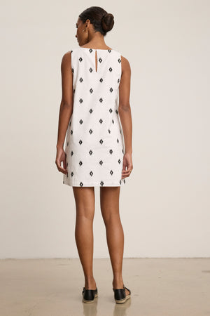 A woman faces away, wearing the ELARA Embroidered Cotton Voile Tank Mini Dress by Velvet by Graham & Spencer—sleeveless, white with black diamond patterns—and black slip-on shoes against a plain light background.