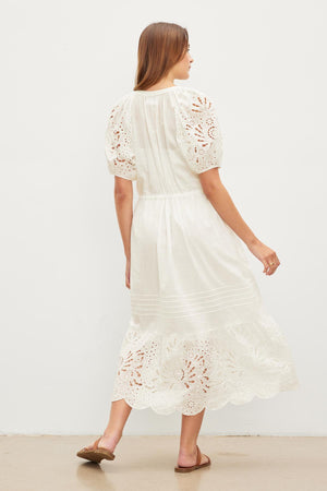 A woman standing facing away from the camera, wearing a white NADIA EMBROIDERED COTTON LACE DRESS with puffed sleeves, paired with brown flats.