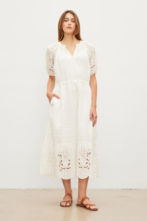 A woman standing in a white room wearing a white, NADIA EMBROIDERED COTTON LACE DRESS with short sleeves and brown sandals. She has shoulder-length brown hair and a neutral expression.
