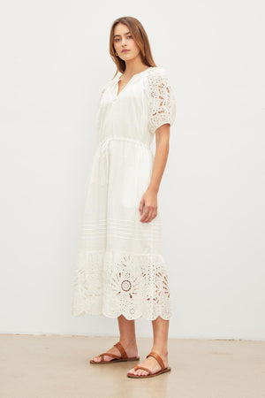 Woman in a white NADIA EMBROIDERED COTTON LACE DRESS with puff sleeves and brown sandals standing against a plain background. (Brand Name: Velvet by Graham & Spencer)