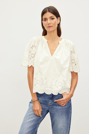 A woman in a white RAZI EMBROIDERED COTTON LACE TOP with scallop cuffs and hem, and blue jeans standing against a plain background by Velvet by Graham & Spencer.
