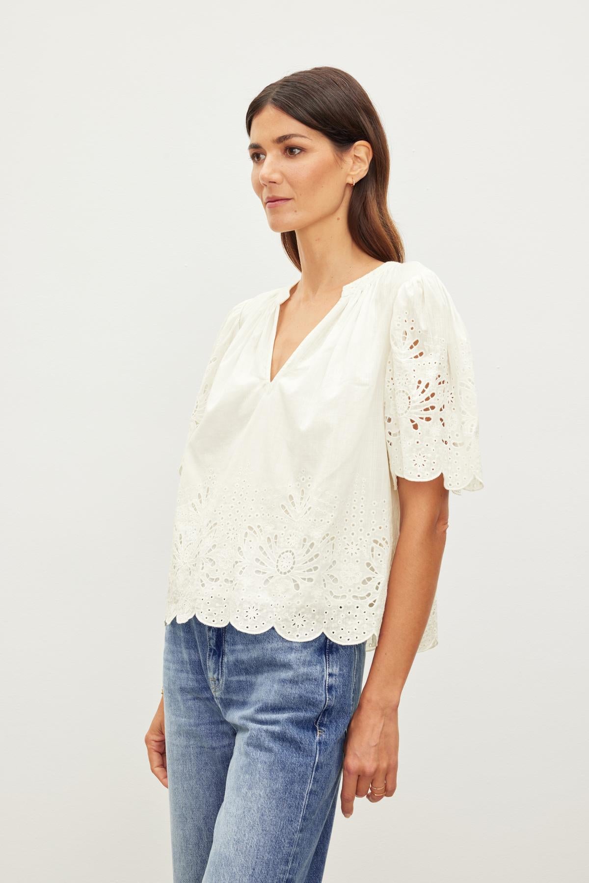   Woman in a white Velvet by Graham & Spencer RAZI EMBROIDERED COTTON LACE TOP with scallop cuffs and hem, and blue jeans standing against a neutral background, looking to the side. 