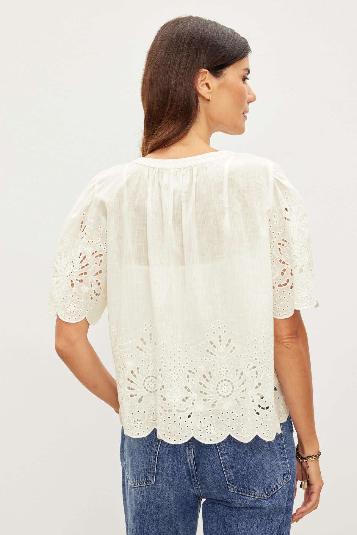   Woman in a white blouse with RAZI EMBROIDERED COTTON LACE details, viewed from the back, standing against a plain background. 