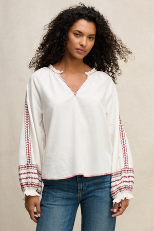 A woman wears the SCARLETT TOP by Velvet by Graham & Spencer, a loose-fitting cotton poplin blouse with red embroidery on the sleeves and cuffs, paired with blue jeans for effortless elegance against a neutral background.