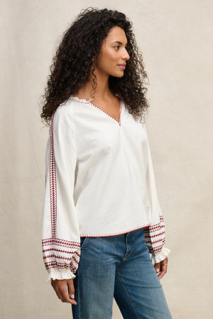 A woman stands sideways against a neutral background, wearing the SCARLETT TOP by Velvet by Graham & Spencer—a white blouse with red embroidery on the sleeves and neckline—paired with blue jeans.