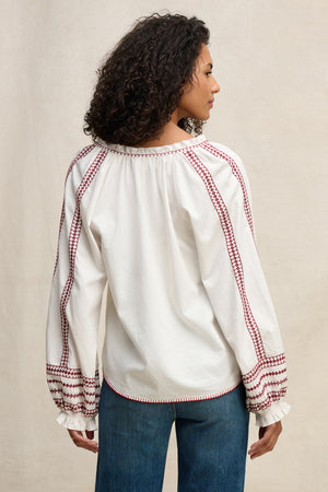 A woman stands with her back turned, wearing the SCARLETT TOP by Velvet by Graham & Spencer—a white embroidered blouse with red details—paired with blue jeans against a plain beige background.