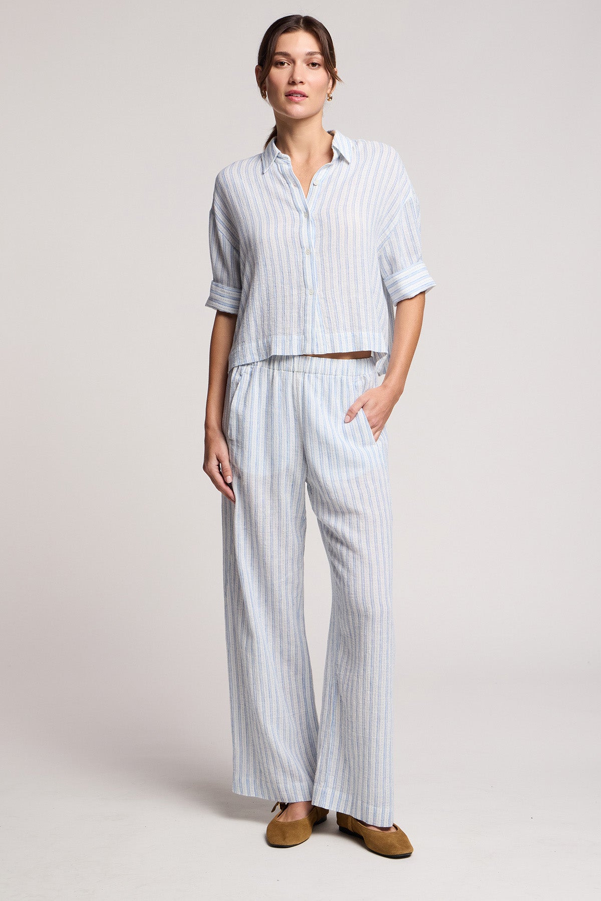 A woman wears the GREYA SHIRT by Velvet by Graham & Spencer, a loose-fitting light blue and white cotton-linen blend summer shirt with embroidered stripes, paired with matching wide-leg pants and brown flats.-41645018022081