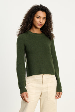 A woman wears the SAVANNAH SWEATER by Velvet by Graham & Spencer, a dark green wool blend, paired with beige pants against a plain white background.