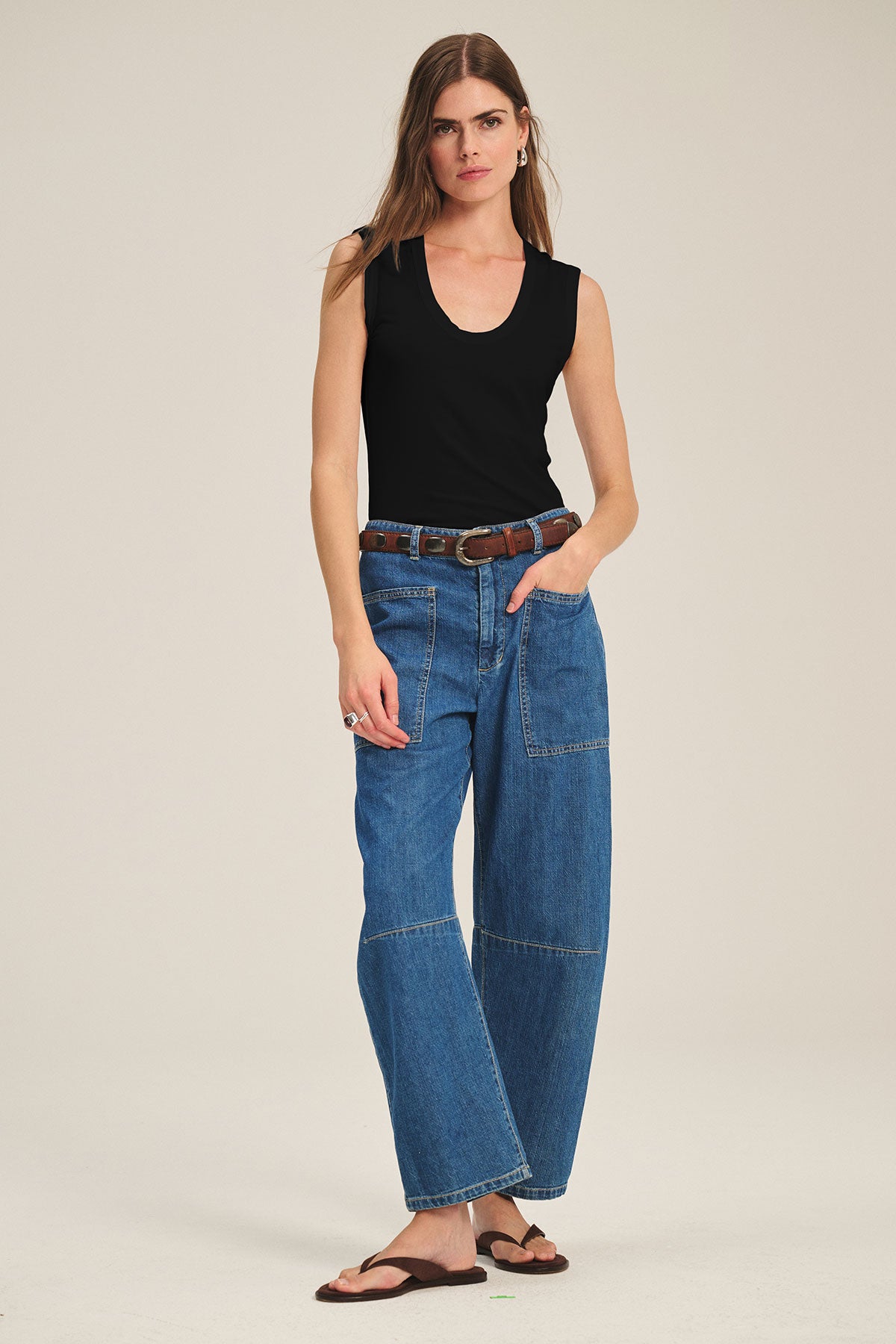 A woman stands against a plain background wearing the ESTINA TANK TOP by Velvet by Graham & Spencer, wide-leg blue jeans with front pockets, a brown belt, and brown sandals.