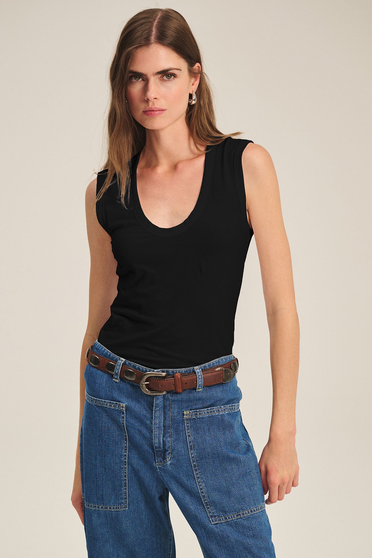 A woman faces forward against a plain light background, wearing the ESTINA TANK TOP by Velvet by Graham & Spencer in black, paired with blue jeans featuring large pockets, a brown belt, and small earrings.-40184100061377
