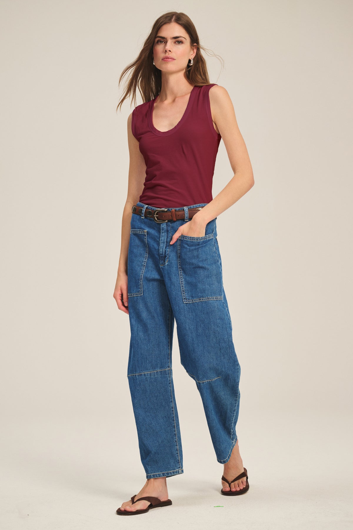A woman stands against a plain background wearing the Velvet by Graham & Spencer ESTINA TANK TOP in maroon, styled with blue patchwork jeans, a brown belt, and black sandals, her left hand tucked into her pocket.-40182510551233