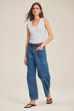 A woman stands against a plain background, wearing the ESTINA TANK TOP by Velvet by Graham & Spencer in light blue, blue patchwork jeans with a brown belt, and dark sandals, one hand casually in her pocket.