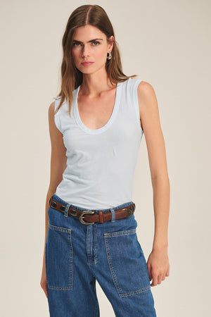 A woman wears the ESTINA TANK TOP by Velvet by Graham & Spencer, blue jeans with front pockets, and a brown belt while standing against a plain background.
