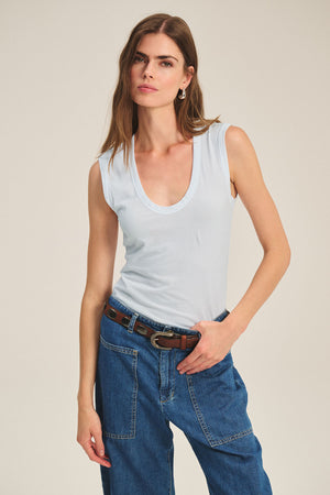 A woman wears the ESTINA TANK TOP by Velvet by Graham & Spencer with blue jeans and a brown belt, standing against a plain background.