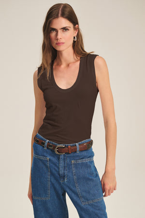A woman stands against a plain background wearing the ESTINA TANK TOP by Velvet by Graham & Spencer, paired with blue jeans featuring patch pockets and a brown belt.