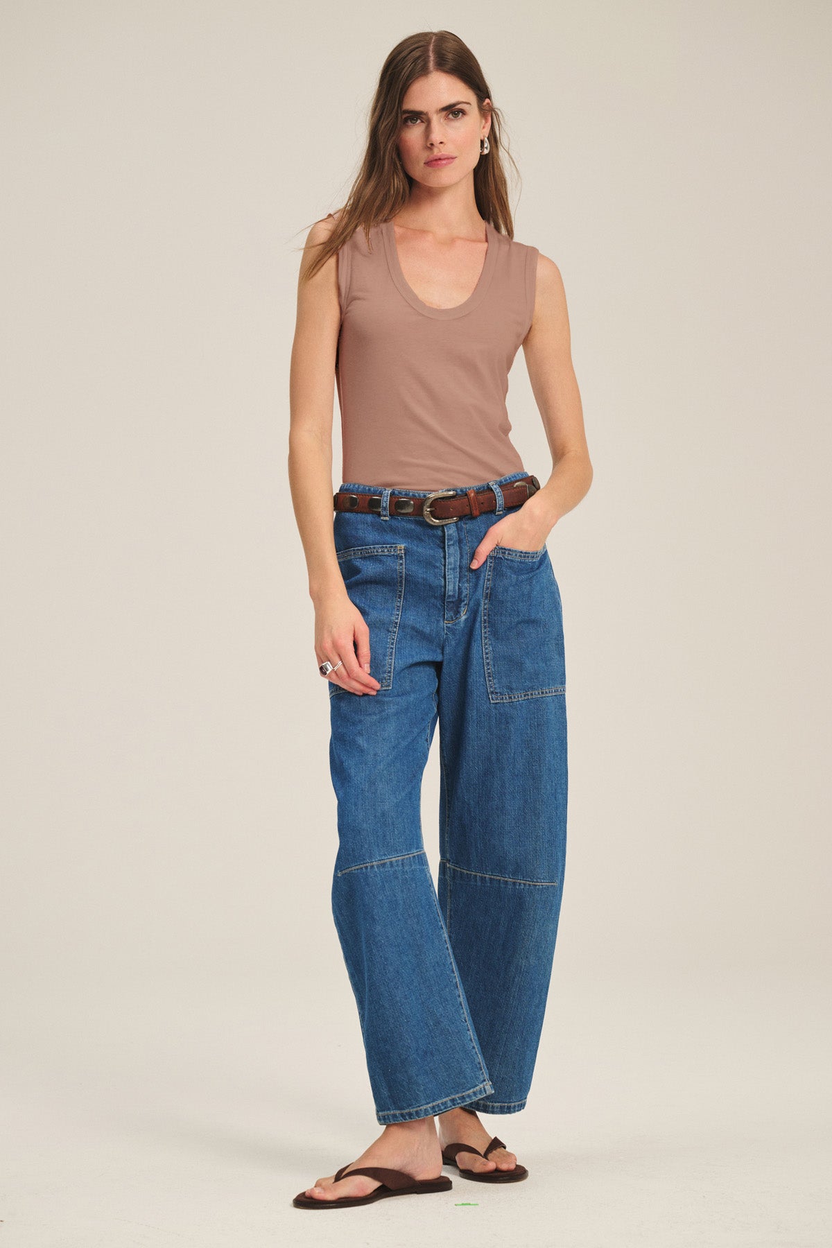 A woman wears the ESTINA TANK TOP by Velvet by Graham & Spencer— a sleeveless beige scoop-neck tank—paired with blue wide-leg jeans, a brown belt, and brown sandals against a plain background.-41637036589249
