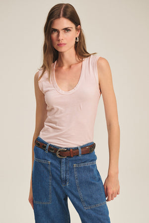 A woman wears the ESTINA TANK TOP by Velvet by Graham & Spencer in pale pink, paired with blue denim jeans featuring visible stitching and a brown belt, standing against a plain background.