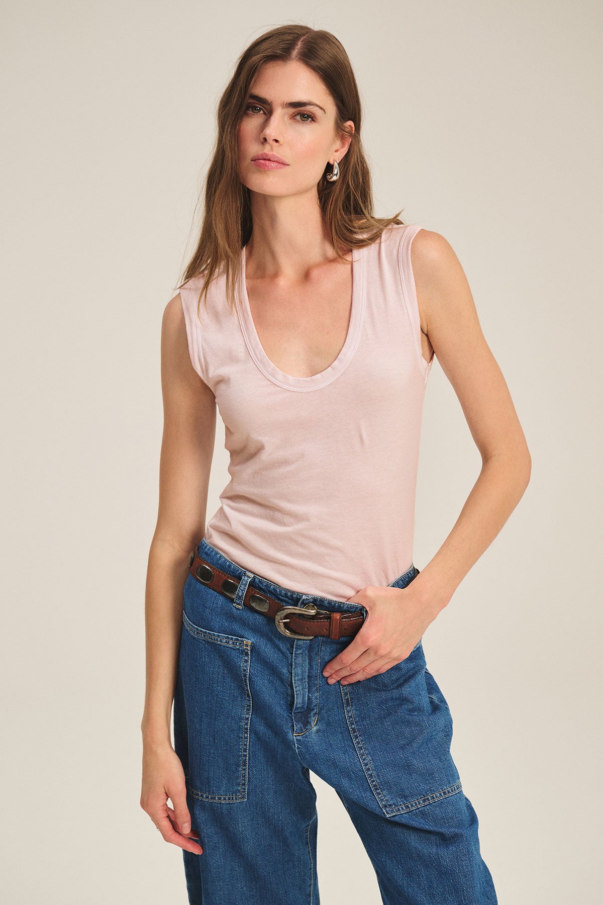 A woman wears the ESTINA TANK TOP by Velvet by Graham & Spencer in light pink, paired with blue jeans and a brown belt, standing against a plain background.-39840854147265