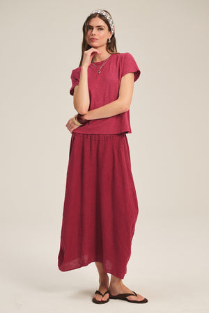 A woman stands against a plain background wearing the Velvet by Graham & Spencer MARIE TEE in dark pink cotton slub with a matching long skirt, black sandals, and a patterned headband.