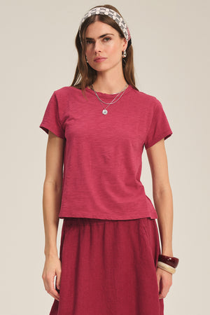 A woman wears the MARIE TEE by Velvet by Graham & Spencer, a relaxed-fit red cotton slub short-sleeve top paired with a matching skirt and accessorized with jewelry and a headscarf, posing against a plain background.