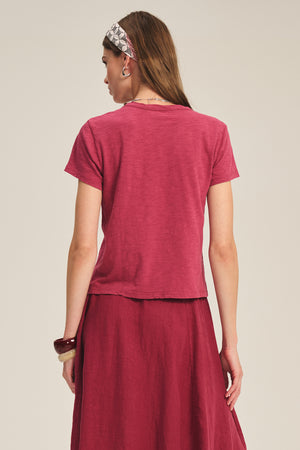 A woman with long hair and a patterned headband wears the Velvet by Graham & Spencer MARIE TEE, a relaxed fit, short-sleeve red cotton slub top, paired with a matching skirt, standing with her back to the camera against a plain background.