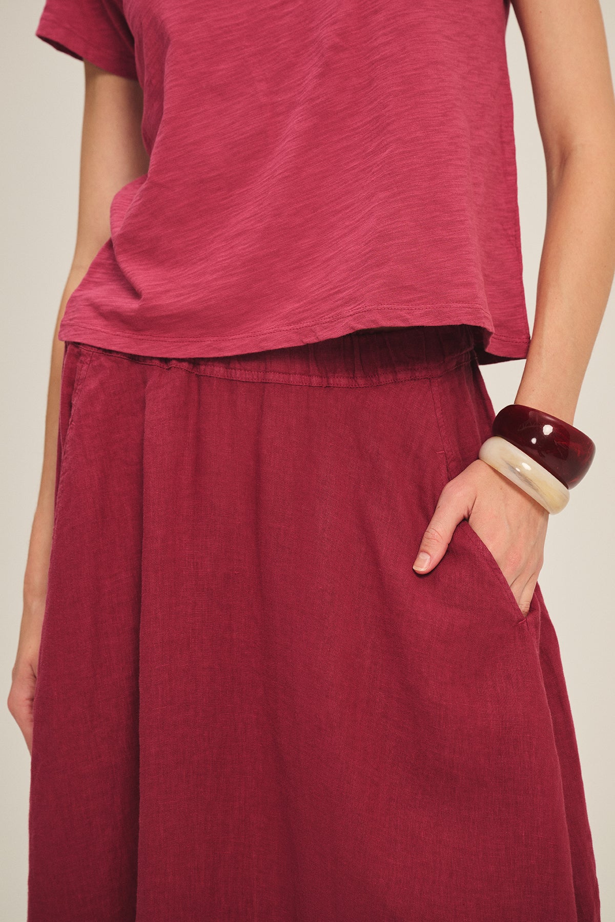 A person wears the Velvet by Graham & Spencer MARIE TEE in red—relaxed fit, short sleeves—paired with a matching red skirt, one hand in the pocket, and accessorized with two chunky bangles.