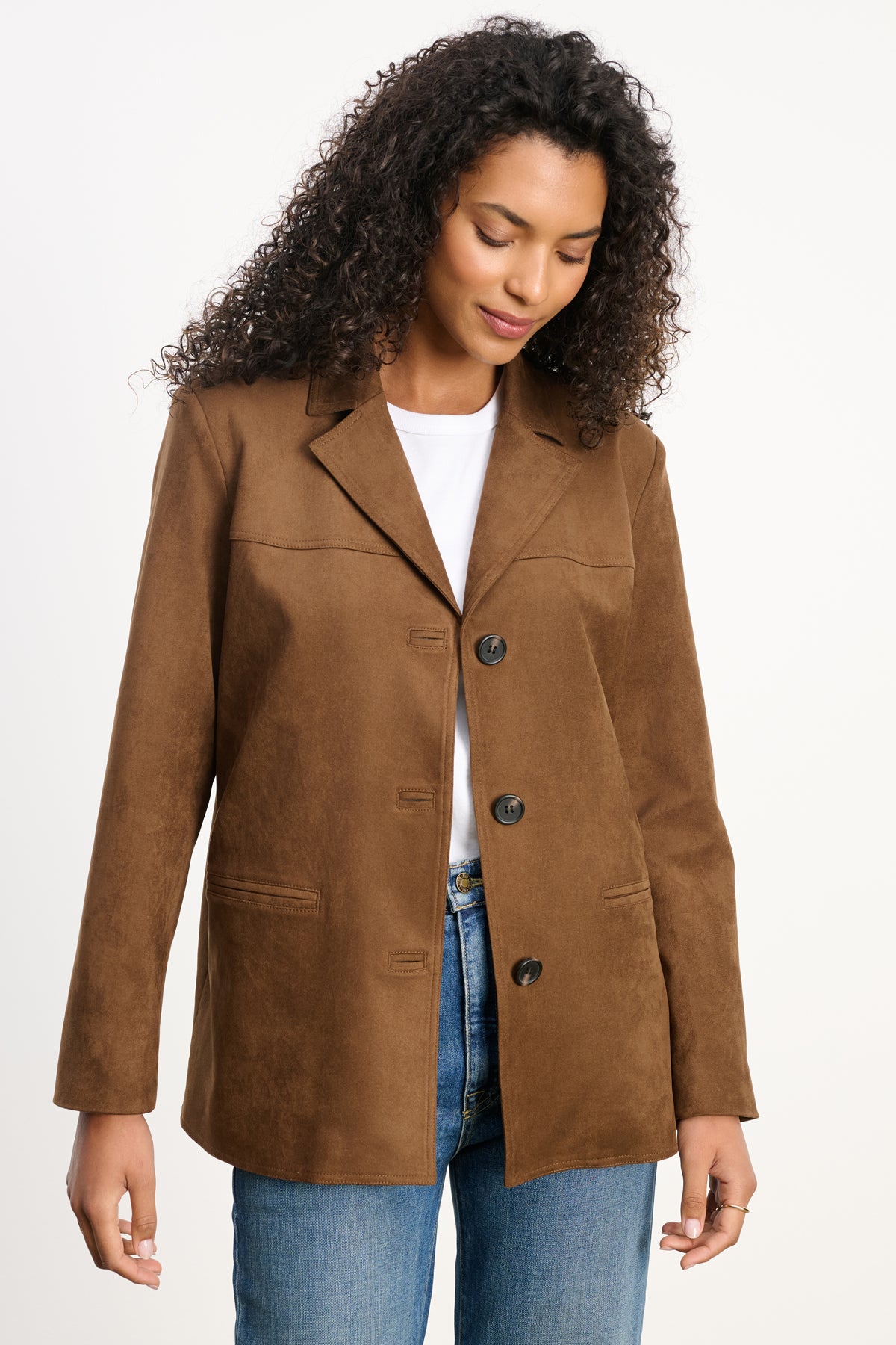 A woman wears the SALIE FAUX SUEDE JACKET by Velvet by Graham & Spencer over a white shirt and blue jeans, standing against a plain background while looking down and smiling slightly.-39742393024705