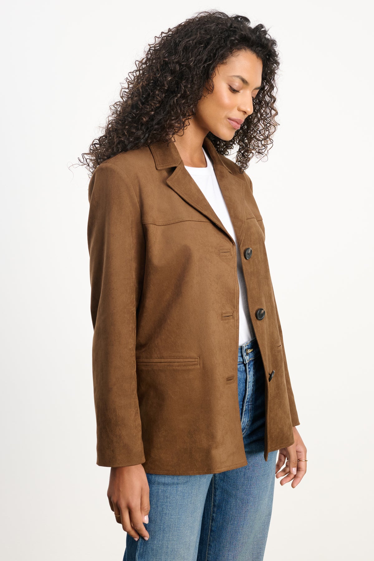 A woman wearing the SALIE FAUX SUEDE JACKET by Velvet by Graham & Spencer over a white top and blue jeans stands against a plain white background, looking downward.-39742392959169