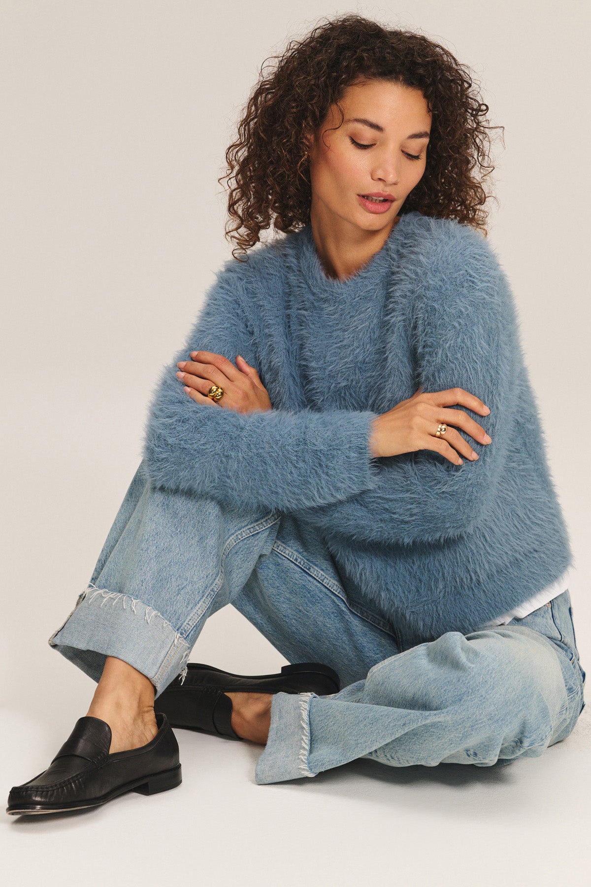 A woman with curly hair sits on the floor with arms crossed, wearing the PENNY SWEATER by Velvet by Graham & Spencer—a blue fuzzy piece crafted from lightweight feather yarn—paired with light blue jeans and black loafers against a neutral background.-40248224612545
