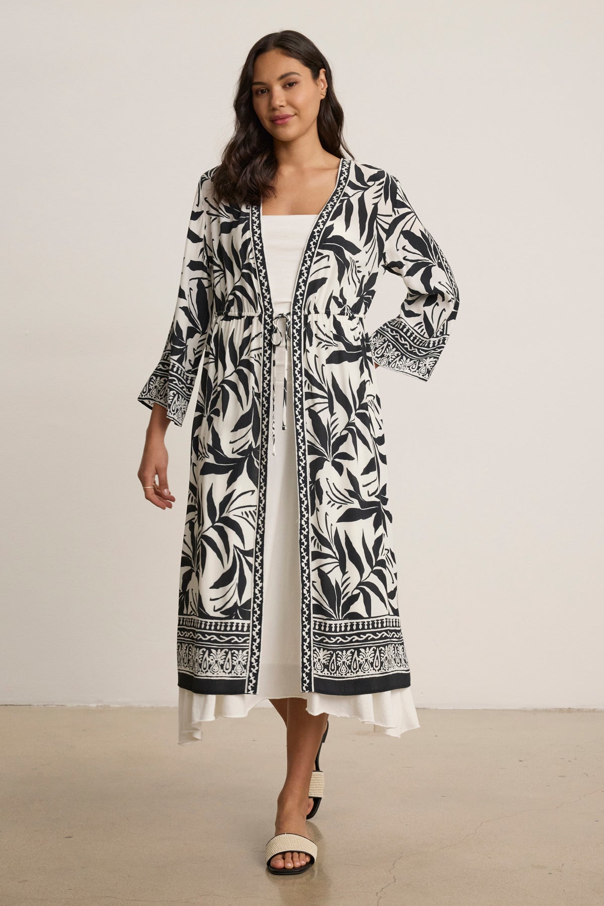 A woman stands indoors wearing the ADDISON COVER UP DRESS by Velvet by Graham & Spencer—a long black and white patterned coat with an adjustable front tie, styled over a white dress and black and white sandals.