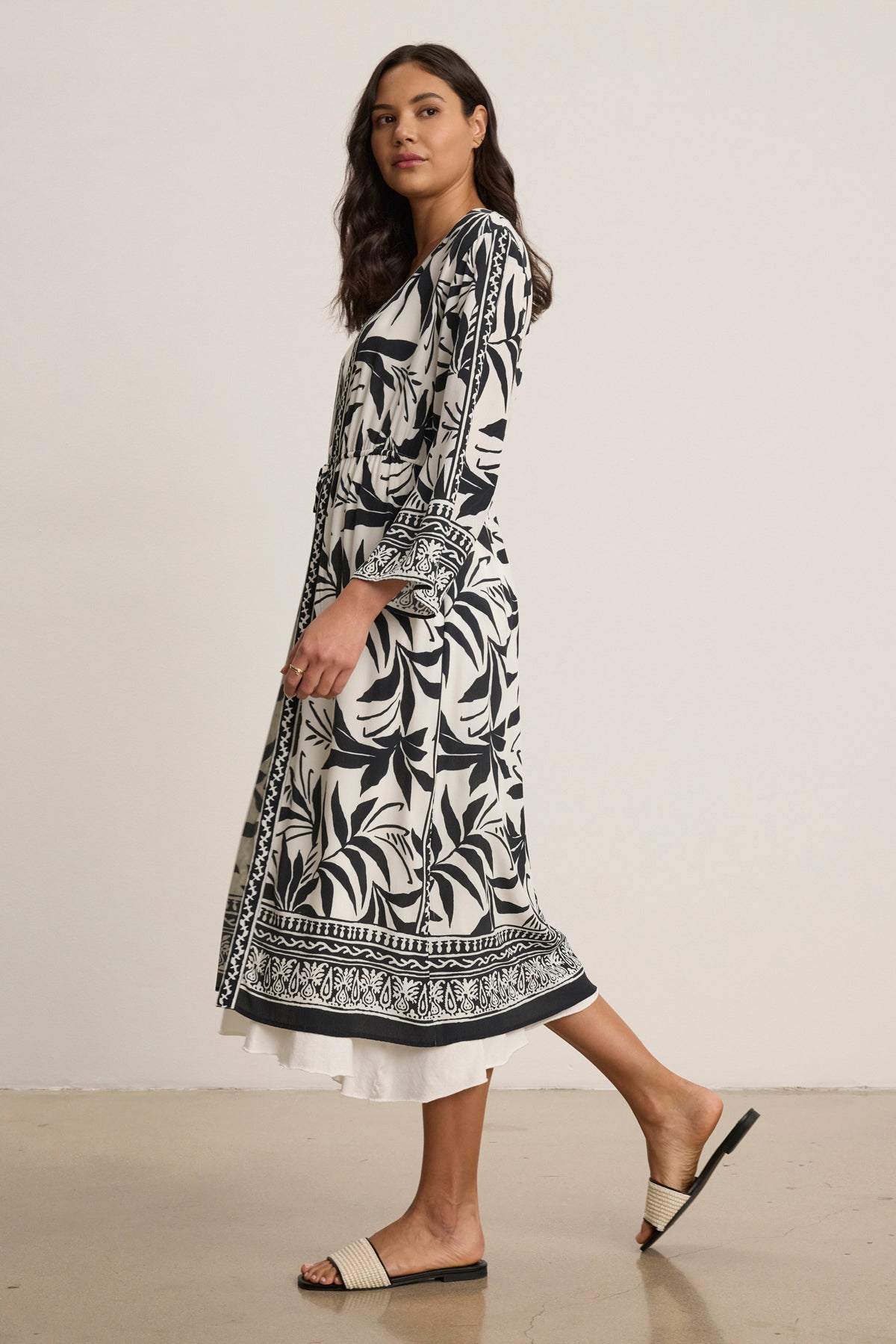 A woman stands sideways wearing the ADDISON COVER UP DRESS by Velvet by Graham & Spencer, a long black and white caftan with long sleeves, an adjustable front tie belt, and flat slide sandals.