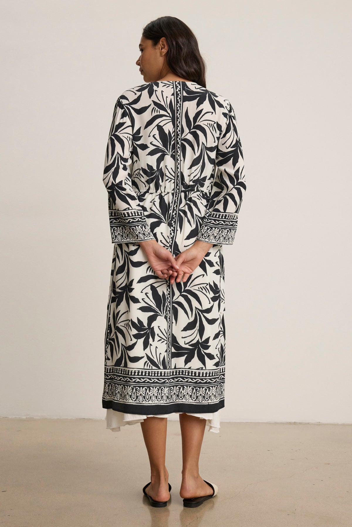A woman faces away, wearing the ADDISON COVER UP DRESS by Velvet by Graham & Spencer—a long-sleeve, black and white patterned caftan—with hands clasped behind her back and black sandals.