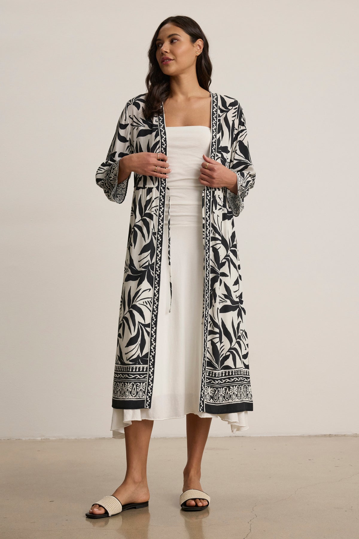 A woman stands indoors wearing the ADDISON COVER UP DRESS by Velvet by Graham & Spencer—a chic, black-and-white patterned caftan layered over a white strapless dress, styled with open-toe sandals.