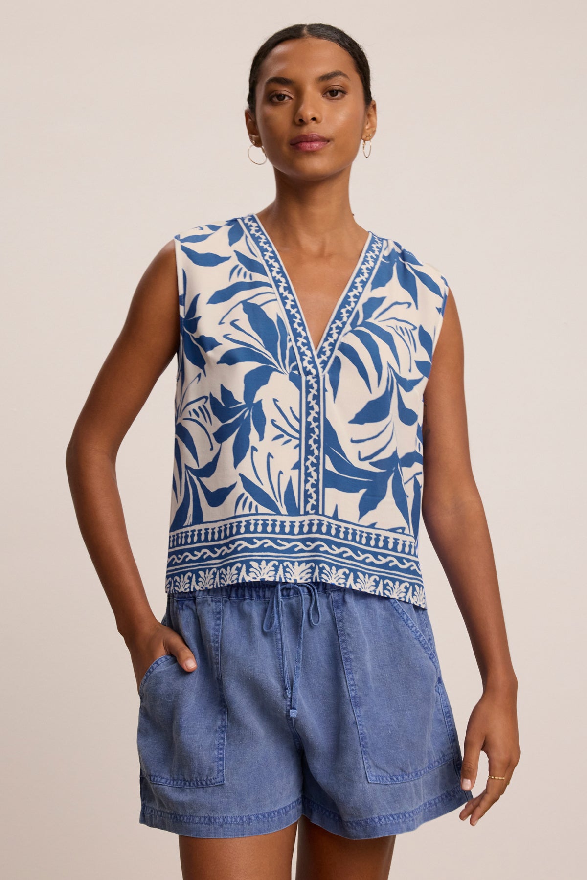 A woman wears the LINDSAY TOP by Velvet by Graham & Spencer, a sleeveless V-neck with blue and white leaf patterns, paired with blue drawstring shorts, standing against a plain background.-39333497503937
