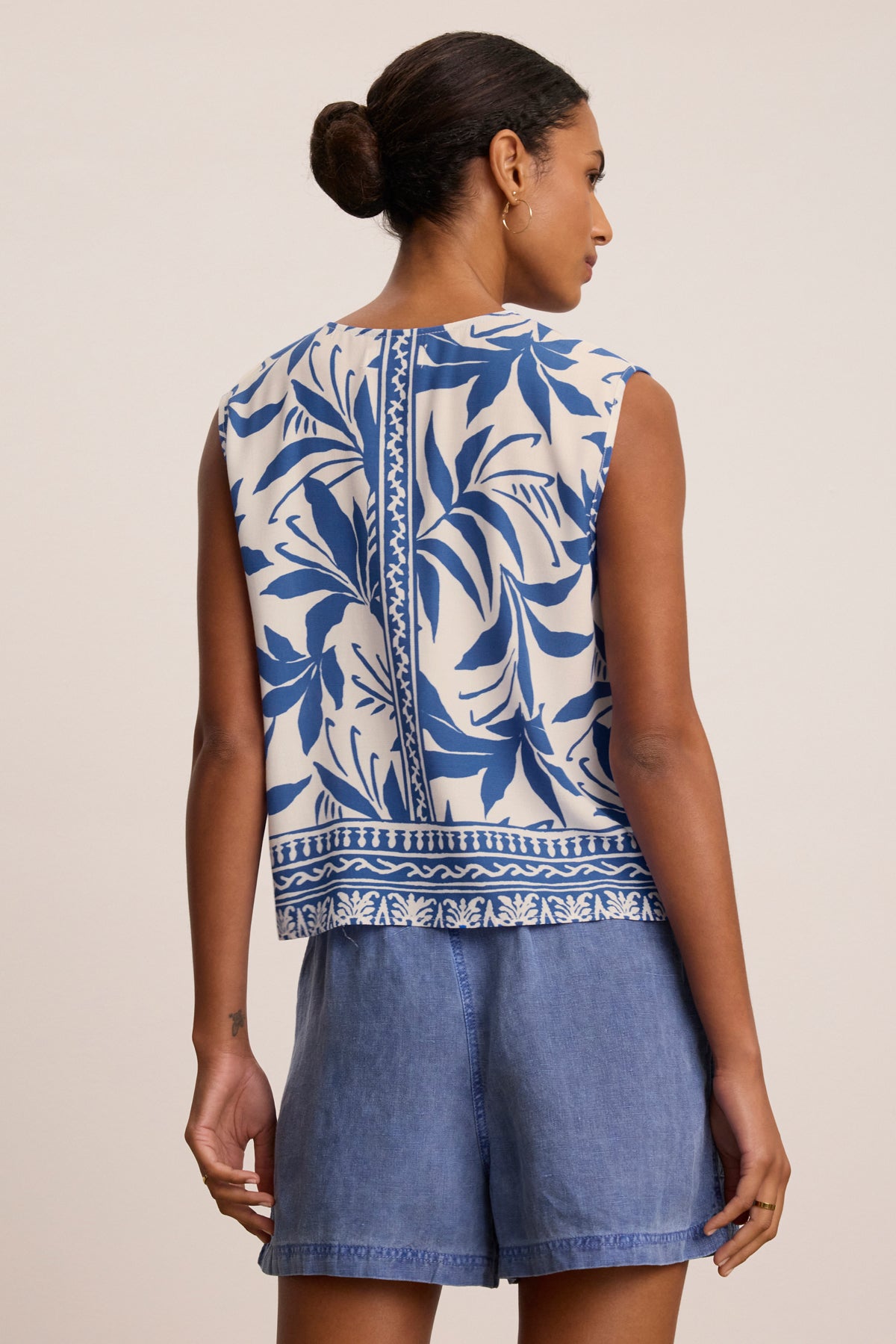 A woman is seen from the back wearing the LINDSAY TOP by Velvet by Graham & Spencer, a lightweight viscose sleeveless top with a blue leaf pattern, paired with blue denim shorts.-39333497569473