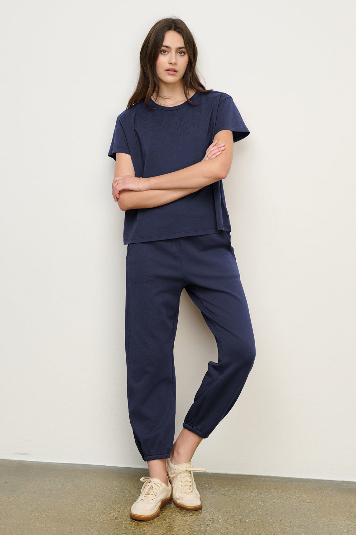 A woman stands against a white wall wearing the Velvet by Jenny Graham ZUMA SWEATPANT in navy with a matching short-sleeve top and beige sneakers.