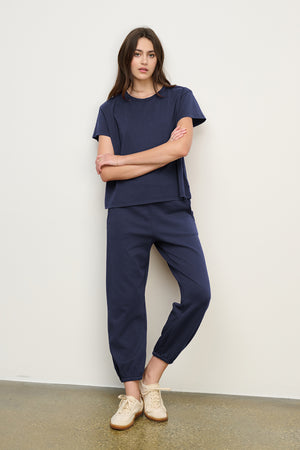 A woman stands against a white wall wearing the Velvet by Jenny Graham ZUMA SWEATPANT in navy with a matching short-sleeve top and beige sneakers.