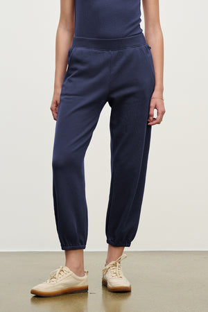 Dressed in Velvet by Jenny Graham's ZUMA SWEATPANT in navy blue with a matching top and beige sneakers, a person stands against a plain background.