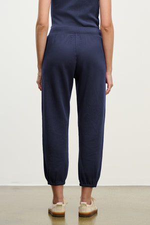 A person stands indoors facing away, wearing Velvet by Jenny Graham's ZUMA SWEATPANT in navy blue with a matching top and beige shoes.