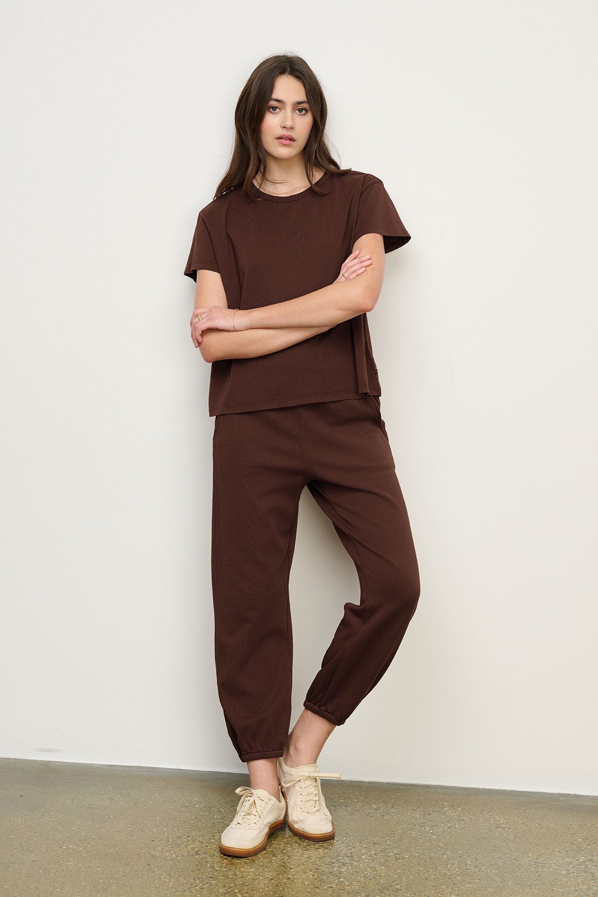 A woman stands against a white wall wearing a relaxed fit brown cotton fleece short-sleeve top and Velvet by Graham & Spencer's ZARA SWEATPANT set with beige sneakers, her arms crossed.-40416936231105
