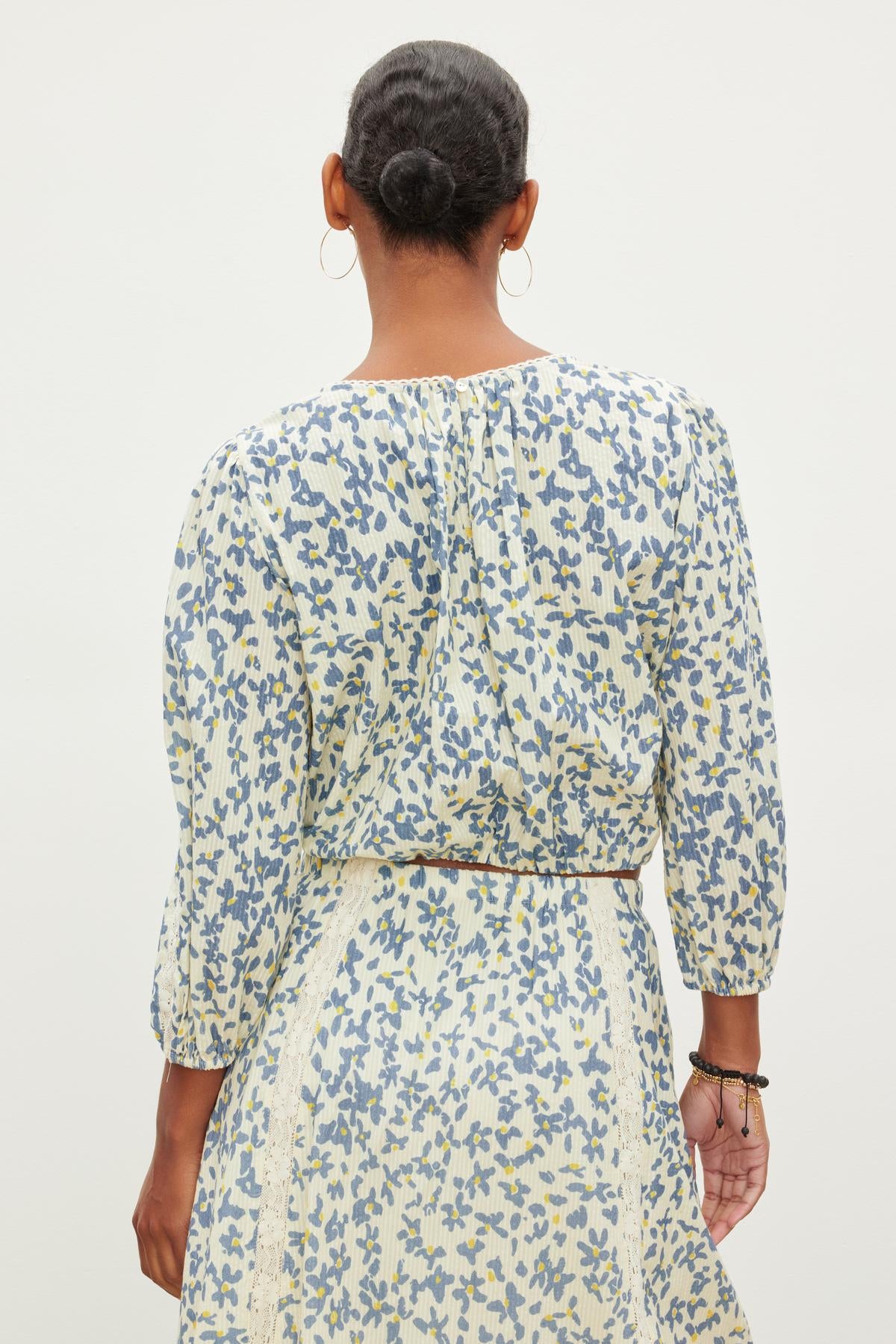 Back view of a person with a bun hairstyle, wearing hoop earrings and a patterned outfit consisting of the CAMERON TOP by Velvet by Graham & Spencer and a skirt in light colors with blue and yellow designs.-37073697800385