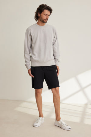 A man stands indoors in a relaxed fit light gray French terry sweatshirt, BOSLOW SHORT black drawstring shorts by Velvet by Graham & Spencer, and white sneakers, against a neutral background with sunlight streaming in.