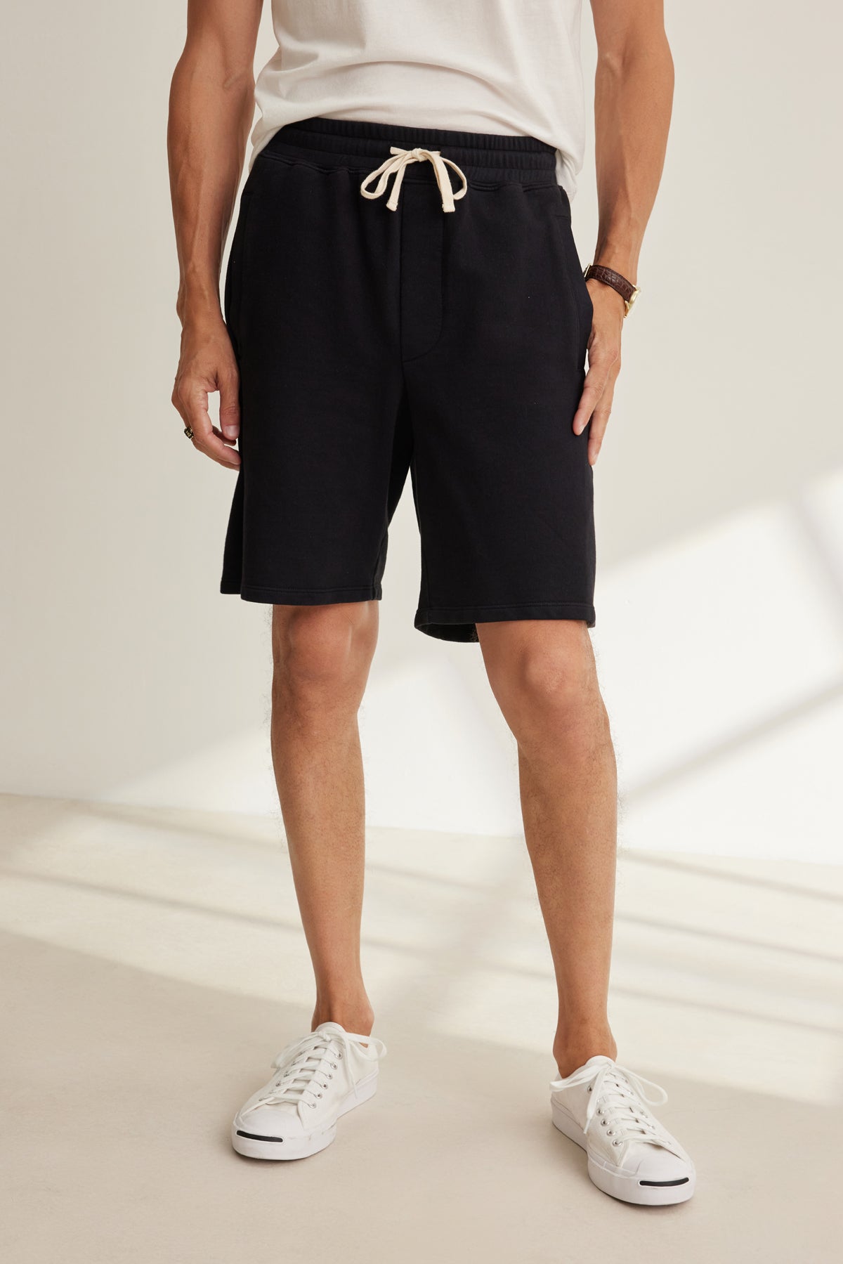 A person wearing a white t-shirt, Velvet by Graham & Spencer BOSLOW SHORT black drawstring shorts, white sneakers, and a brown bracelet stands indoors on a light-colored floor.