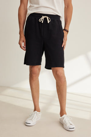 A person wearing a white t-shirt, Velvet by Graham & Spencer BOSLOW SHORT black drawstring shorts, white sneakers, and a brown bracelet stands indoors on a light-colored floor.