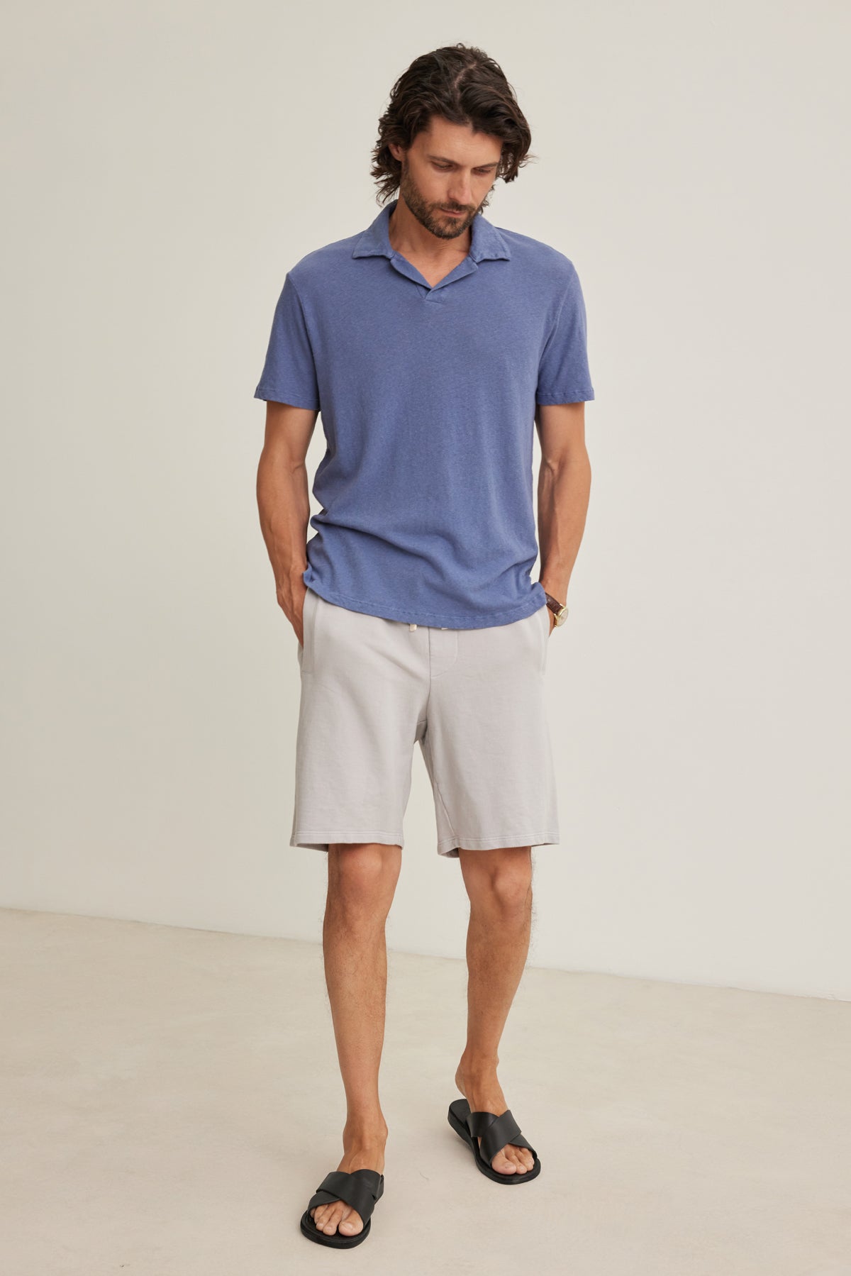 A man stands indoors with hands in his pockets, wearing a blue polo, black slides, and Velvet by Graham & Spencer BOSLOW SHORT relaxed fit light gray shorts featuring a drawstring elastic waist.
