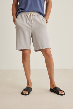 A person wearing the Velvet by Graham & Spencer BOSLOW SHORT—a relaxed fit, light gray French terry short with a drawstring waist—stands with hands in pockets, paired with a blue shirt and black slide sandals against a plain background.